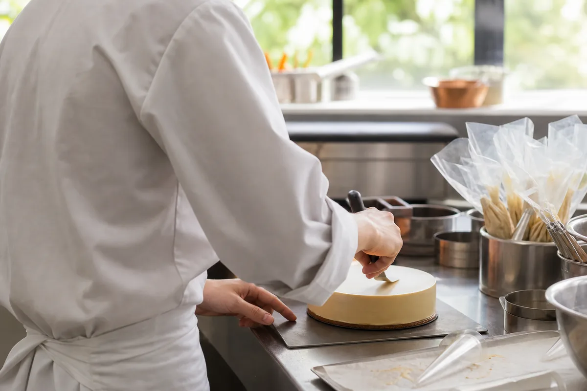 Jeune apprenti en veste blanche dressant un entremets à la spatule dans un laboratoire de pâtisserie lumineux